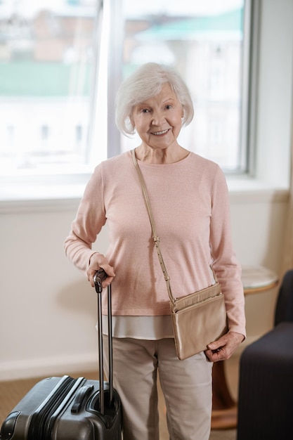 Belle femme senior debout avec une valise dans une chambre d'hôtel