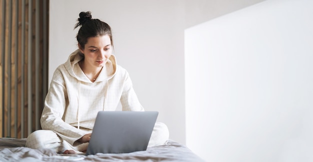 Belle femme souriante adolescente étudiante pigiste aux cheveux longs noirs en décontracté à l'aide d'un ordinateur portable assis sur le lit à la bannière de la maison