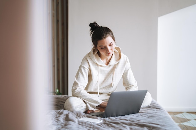 Belle femme souriante adolescente étudiante pigiste aux cheveux longs noirs en décontracté à l'aide d'un ordinateur portable assis sur le lit à la maison