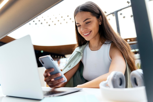Belle femme souriante à l'aide d'un ordinateur portable tenant un smartphone et faisant des achats en ligne sur l'application mobile