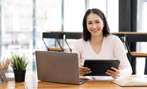 Belle femme souriante à l'aide d'une tablette et d'un ordinateur portable assis à table au bureau souriant à la caméra