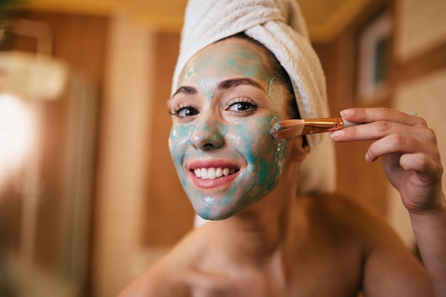 Belle femme souriante appliquant un masque facial dans la salle de bain