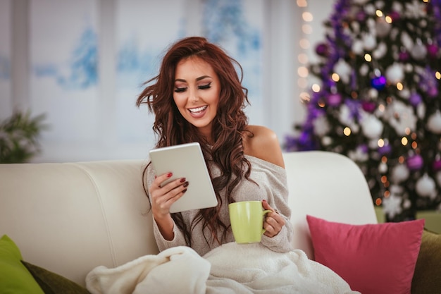 Belle femme souriante assise enveloppée dans une couverture la veille de Noël avec une tasse de thé et à l'aide d'une tablette.