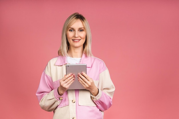 Une belle femme souriante aux cheveux blonds utilise une tablette pour l'éducation ou le travail de cartographie commerciale. Fille positive souriante utilisant des technologies modernes isolées sur fond rose.