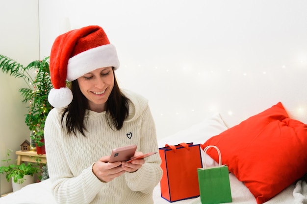 Belle femme souriante en bonnet de noel tenant téléphone et carte bancaire en plastique