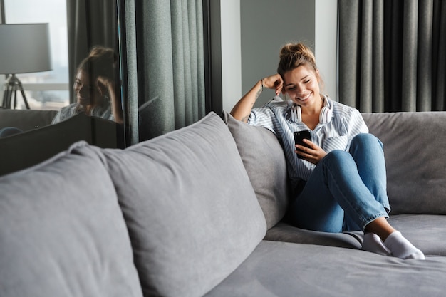 belle femme souriante avec un casque à l'aide d'un téléphone portable assis sur un canapé dans le salon