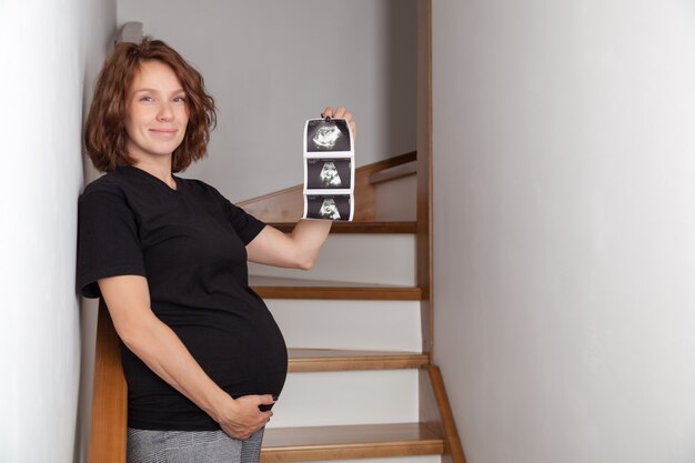 Belle femme souriante caucasienne bouclée enceinte assise sur des escaliers en bois et montrant des échographies