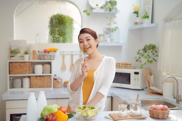 Belle femme souriante mangeant une salade végétarienne biologique fraîche dans une cuisine moderne.