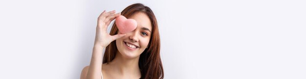 Belle femme souriante avec un maquillage naturel de peau propre et des dents blanches sur fond gris tenant un coeur hygiénique rose