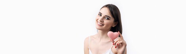 Belle femme souriante avec un maquillage naturel de peau propre et des dents blanches sur fond gris tenant un coeur hygiénique rose