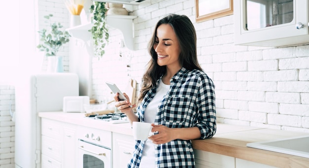 Belle femme souriante mignonne utilise un téléphone intelligent dans la cuisine à la maison