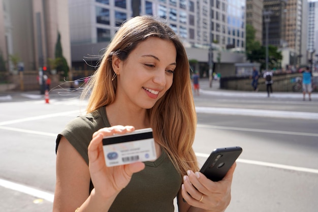 Belle femme souriante montrant une carte de crédit en regardant son téléphone à l'extérieur