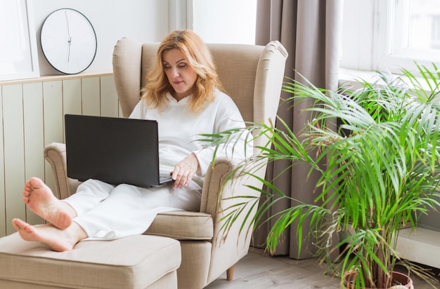 Belle femme souriante mûre travaillant sur un ordinateur portable et assise dans une grande chaise confortable à la maison