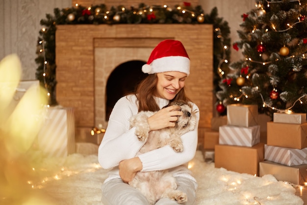 Belle femme souriante portant un pull blanc et un chapeau de père Noël rouge jouant avec un chien pékinois dans un salon festif près de la cheminée et de l'arbre de Noël, assise sur le sol sur un tapis moelleux.