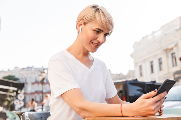 Belle femme souriante portant un t-shirt blanc à l'aide d'un téléphone portable et d'un écouteur sans fil, assis dans un café à l'extérieur en été et buvant du café dans une tasse en papier