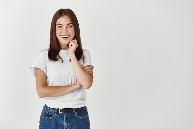 Belle femme souriante et regardant avec intérêt devant, vérifiant quelque chose d'intéressant, debout sur un mur blanc.