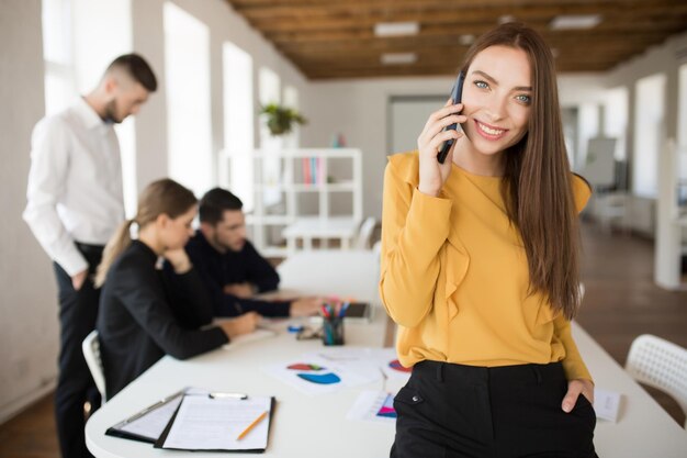 Belle femme souriante regardant rêveusement à huis clos tout en parlant au téléphone portable au bureau avec des collègues en arrière-plan