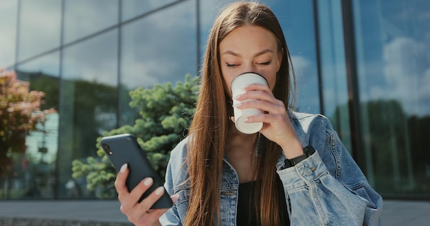 Belle femme souriante tapant un message près de l'immeuble de bureaux pendant la pause-café Fille utilisant un smartphone et buvant du café sur le banc de la rue