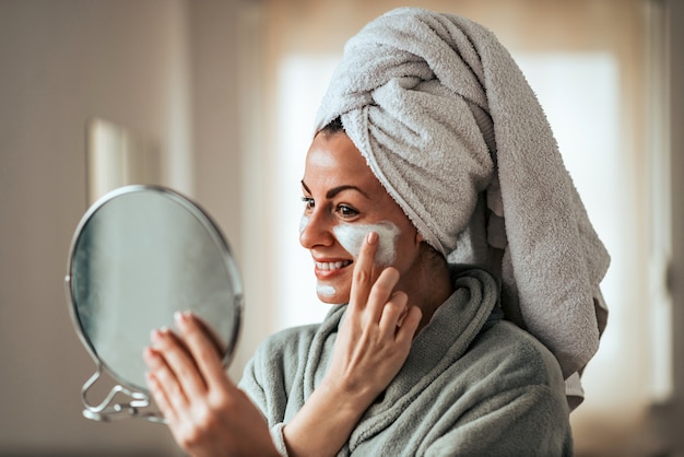 Belle femme souriante tenant un miroir et appliquant une crème pour le visage à la maison.