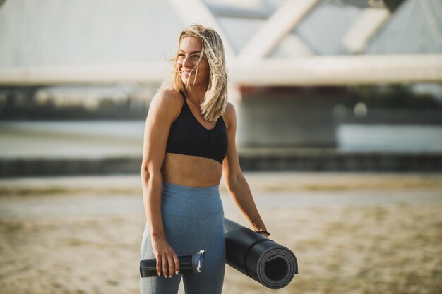 Belle femme souriante en vêtements de sport tenant un tapis de yoga et une bouteille d'eau et marchant avant de s'entraîner près de la rivière dans la ville.