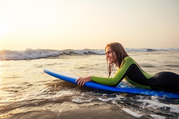 Belle femme sportive en combinaison de plongée allongée sur une planche de surf en attente d'une grosse vague. Fille de surf en combinaison surfant dans l'océan au coucher du soleil. Cheveux mouillés, bonheur et liberté vacances à la plage