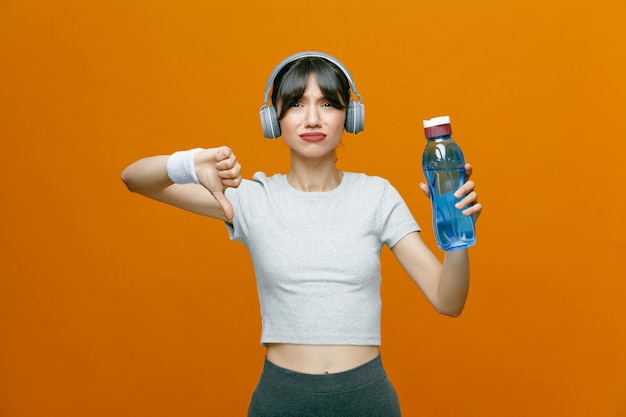 Belle femme sportive en tenue de sport avec un casque tenant une bouteille d'eau regardant la caméra insatisfaite montrant le pouce vers le bas debout sur fond orange