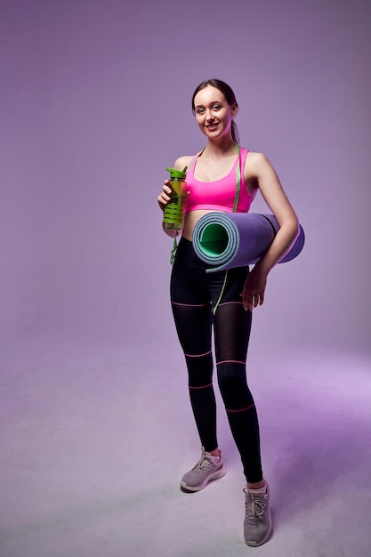 Belle femme sportive en tenue de sport tenant une bouteille d'eau et tapis de fitness isolé sur violet. Aller à la gym.