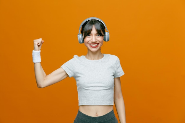 Belle femme sportive en vêtements de sport avec un casque regardant la caméra en souriant confiant levant le poing fermé debout sur fond orange