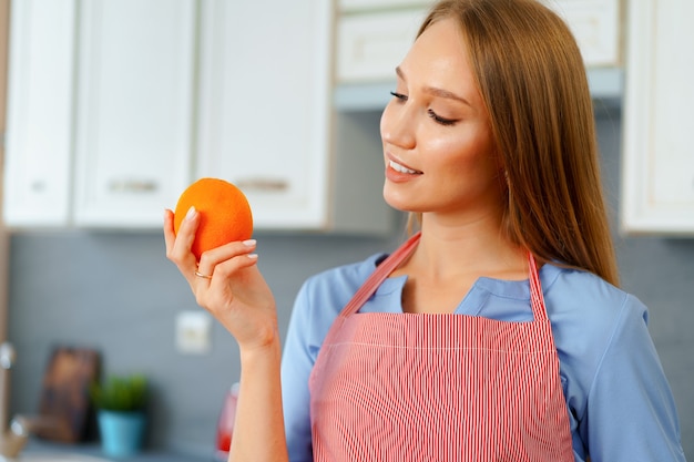 Belle femme en tablier rouge tenant des oranges mûres en se tenant debout dans la cuisine