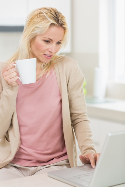 Belle femme avec une tasse de café en utilisant un ordinateur portable dans la cuisine