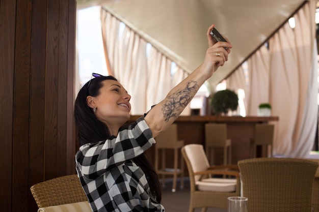 Belle femme avec un tatouage et une chemise à carreaux fait du selfie avec un sourire dans un beau café.