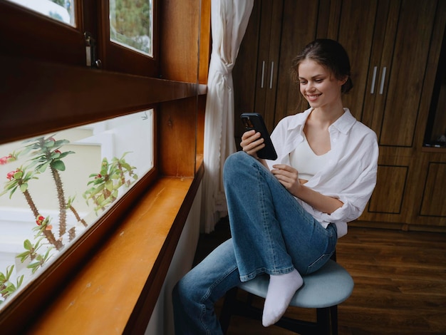Belle femme avec téléphone à la main assise à la fenêtre avec cadre en bois de la maison maison style de vie confortable avec travail en ligne atmosphère confortable et l'esthétique de la chaleur printemps