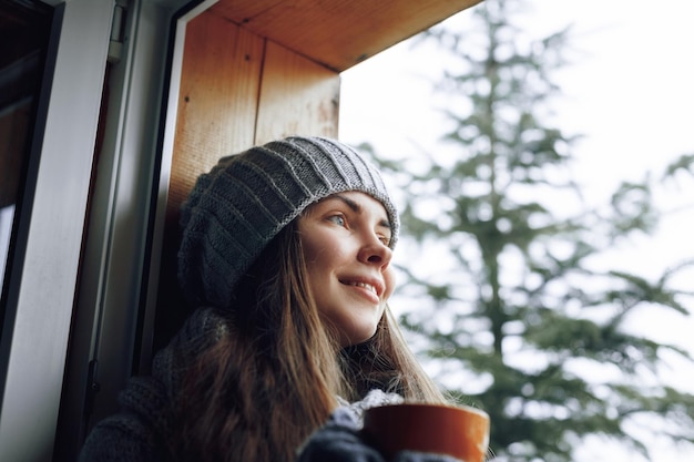 Belle femme tenant et buvant une tasse de café ou de cacao dans des gants assis à la maison près de la fenêtre