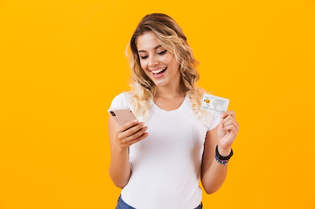 Belle femme tenant un téléphone portable et une carte de crédit en mains, isolé sur un mur jaune