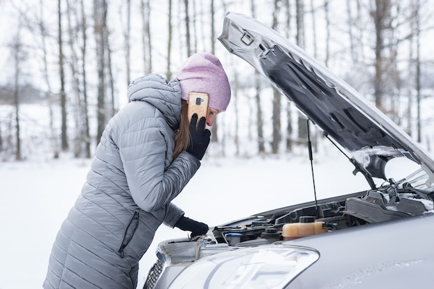 Une belle femme tendue se tient sur la route et attend de l'aide à côté d'une voiture cassée avec le capot ouvert