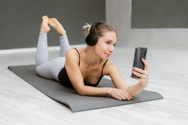 Belle femme en tenue de sport écoutant de la musique avec un casque et utilise un smartphone en position couchée sur un tapis intérieur