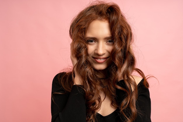 Belle femme tête rouge posant sur un mur rose. Poils ondulés. Sourire parfait.