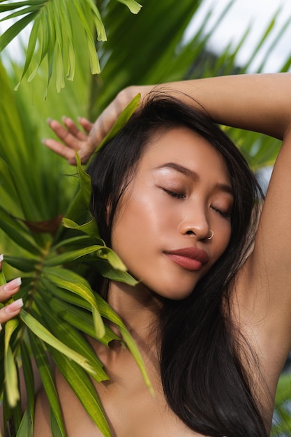 Belle femme thaïlandaise asiatique avec une peau bronzée douce et propre, portrait de beauté, soins de la peau positifs, posant avec des feuilles vertes, des lèvres charnues, une beauté naturelle en plein air