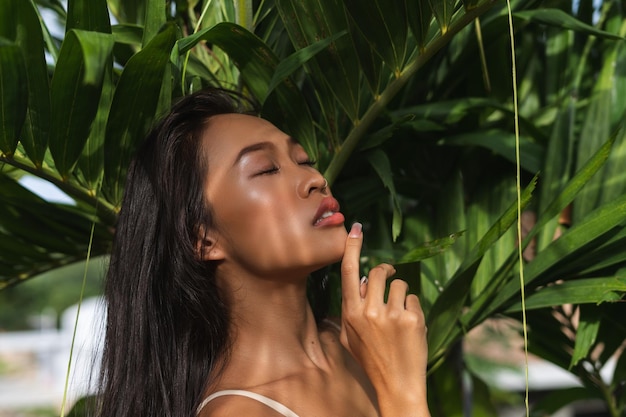 Belle femme thaïlandaise asiatique avec une peau bronzée douce et propre posant par des feuilles de palmier