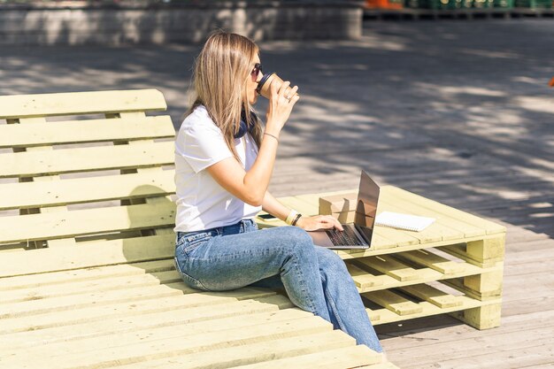 Belle femme travaillant au cahier s'asseoir sur un banc à l'extérieur et boire du café. En utilisant l'ordinateur