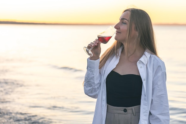 Belle femme avec un verre de vin sur fond de mer au coucher du soleil