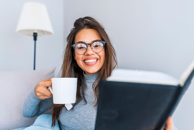 Belle femme en vêtements décontractés et lunettes lit un livre, tenant une tasse