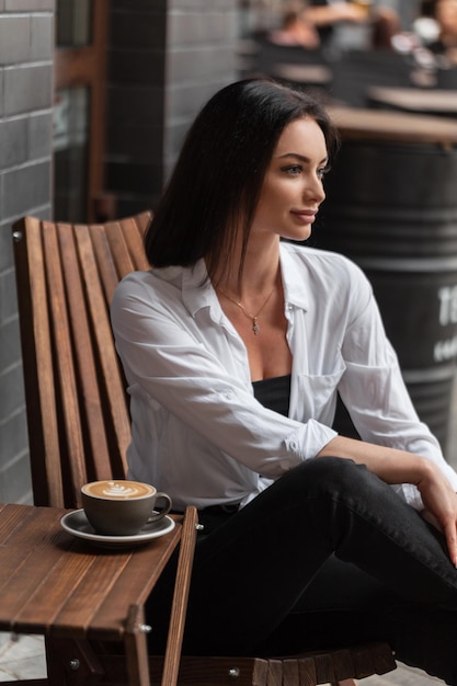 Belle femme en vêtements décontractés de mode avec chemise est assise sur une chaise en bois et boit du café cappuccino dans la rue