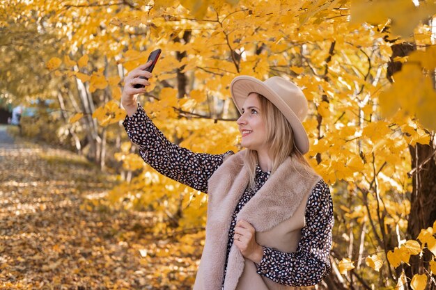 Belle femme en vêtements à la mode et chapeau prend selfie sur téléphone près de l'arbre d'automne jaune.