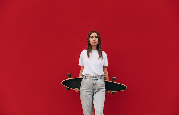 Belle femme en vêtements de rue élégants se dresse sur un fond rouge avec un longboard