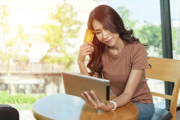 Belle femme vêtue d'une chemise brune assise et souriante, heureuse assise près d'une grande fenêtre, elle fait des achats en ligne avec une tablette et tient une carte de crédit.