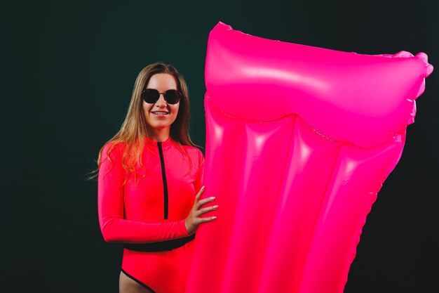 Belle femme vêtue d'un maillot de bain rose et de lunettes de soleil posant avec un matelas gonflable rose isolé sur fond vert