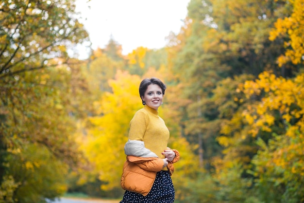 Une belle femme vêtue d'un pull jaune et d'une veste orange sourit et regarde la caméra. Femme appréciant la nature d'automne. photo