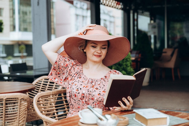 Une belle femme vêtue d'une robe et d'un chapeau lit un livre dans un café de rue femme élégante en robe d'été