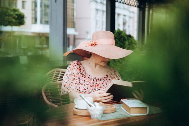 Une belle femme vêtue d'une robe et d'un chapeau lit un livre dans un café de rue femme élégante en robe d'été